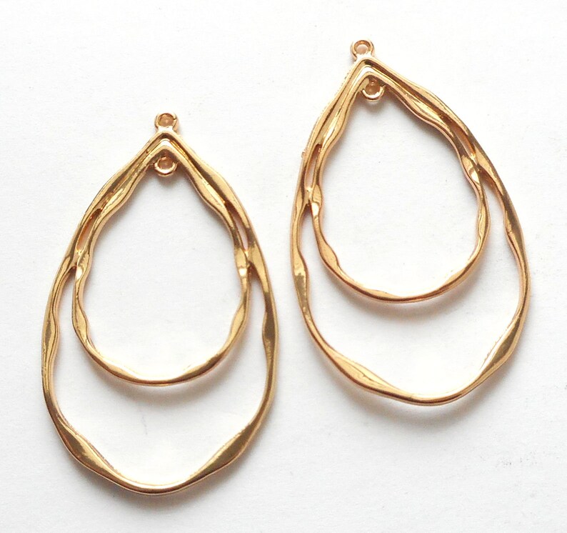 May include: Two gold-colored, teardrop-shaped earring components. Each component features a double-layered design with a textured, wavy outline. The top of each earring has a small loop for attachment. The earrings are set against a white background.
