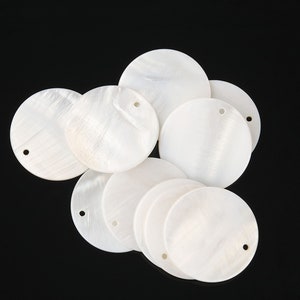 May include: A collection of white, round, mother-of-pearl shell discs with a hole in the centre. The discs are arranged in a circular pattern.