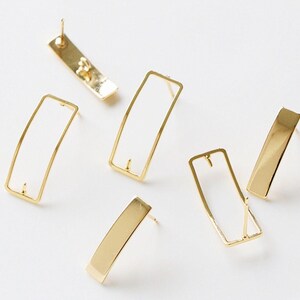 May include: Gold-coloured rectangular stud earrings. The earrings have a solid, rectangular post and a hollow, open rectangular frame. The earrings are displayed on a white surface, highlighting their minimalist design and reflective quality.