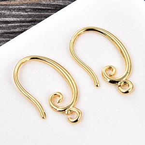May include: Two gold-colored earring hooks. Each hook has a curved, elongated shape with a looped end and a decorative swirl at the bottom. The hooks are displayed on a white surface.