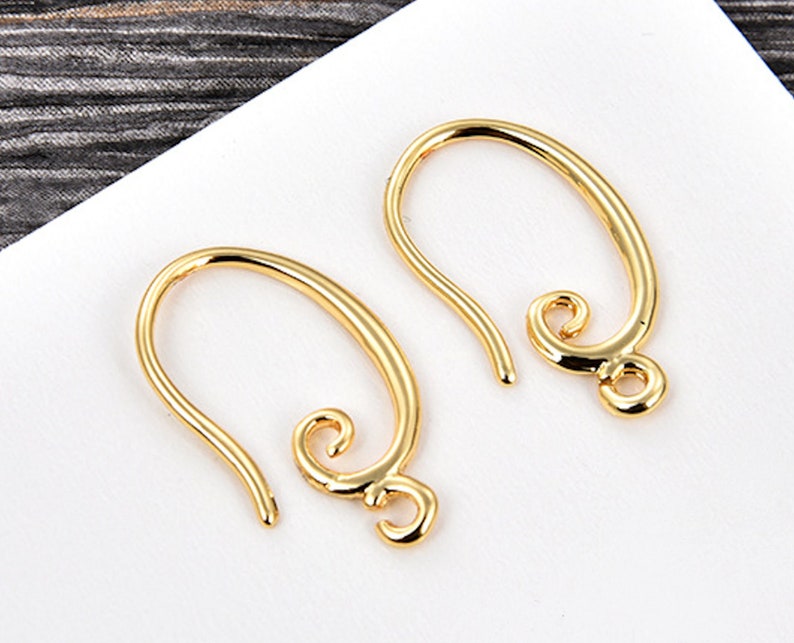 May include: Two gold-colored earring hooks. Each hook has a curved, elongated shape with a looped end and a decorative swirl at the bottom. The hooks are displayed on a white surface.