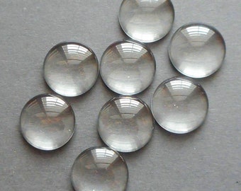 May include: A collection of eight clear, round glass cabochons. Each cabochon is domed and reflects light, creating a shiny appearance. The cabochons are arranged on a gray surface.