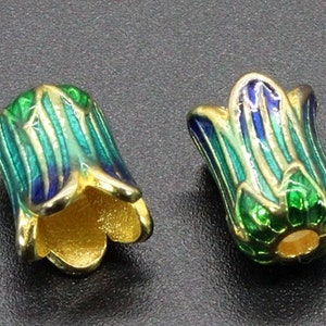May include: Two enameled flower-shaped beads. The beads are gold-colored with green, blue, and turquoise enamel stripes. The beads have a gold-colored interior and are set against a dark gray background. These beads are suitable for jewelry making.