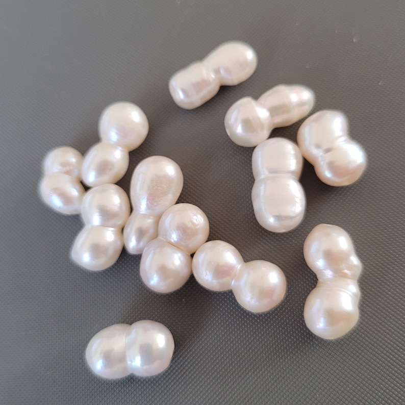 May include: A collection of off-white baroque pearls, each with an irregular shape and a smooth, glossy finish. The pearls are arranged on a gray surface.