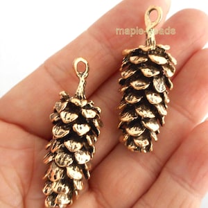 May include: Two gold-tone metal pine cone charms with a loop at the top for hanging. The charms are detailed with textured scales and are approximately 5 cm in length.