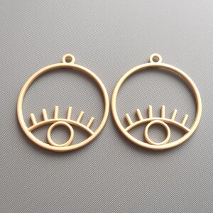May include: A pair of gold-toned earrings with a circle design and an eye shape in the center. The eye has a circle for the pupil and lashes around the top.