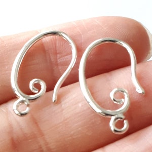 May include: Two silver-colored earring hooks. Each hook has an oval shape with a looped design at the bottom and a decorative swirl at the top. The hooks are designed for jewelry making.
