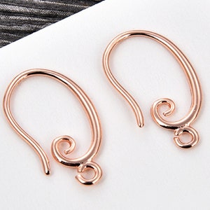 May include: Two rose gold earring hooks. Each hook has a curved, oval shape with a looped end and a decorative swirl at the bottom. The hooks are designed for jewelry making and are displayed against a white background.