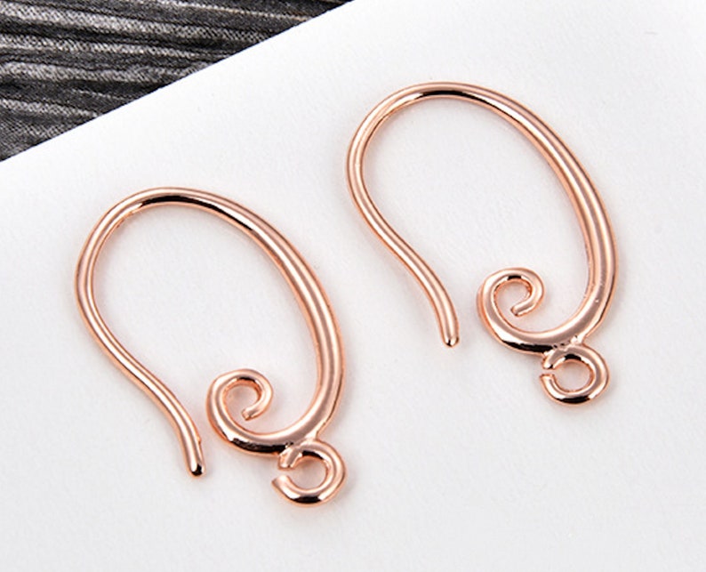 May include: Two rose gold earring hooks. Each hook has a curved, oval shape with a looped end and a decorative swirl at the bottom. The hooks are designed for jewelry making and are displayed against a white background.