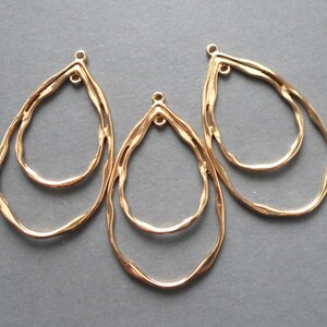 May include: Three gold-colored, teardrop-shaped jewelry findings. Each finding has a double-layered design with a textured, hammered appearance. The top of each finding has a small loop for attaching to other components.