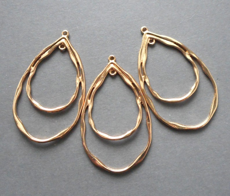 May include: Three gold-colored, teardrop-shaped jewelry findings. Each finding has a double-layered design with a textured, hammered appearance. The top of each finding has a small loop for attaching to other components.