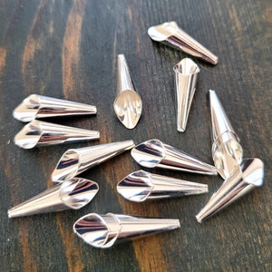 May include: A collection of shiny, silver-colored metal cone-shaped findings. Each piece has a flared, open end and a pointed tip. The findings are scattered on a dark, wooden surface, reflecting light.