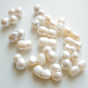 May include: A collection of white baroque pearls, each pearl is a double pearl with two lobes. The pearls are arranged on a white surface.