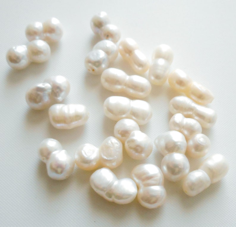 May include: A collection of white baroque pearls, each pearl is a double pearl with two lobes. The pearls are arranged on a white surface.