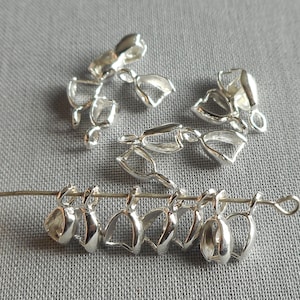 May include: Silver-toned metal pendant bails, a type of jewelry finding, are arranged on a gray surface. Some are strung on a wire, while others are scattered around. These bails are used to attach pendants to necklaces or bracelets.