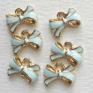 May include: Six gold-toned metal charms shaped like bows with light blue enamel. The bows have a loop at the top for attaching to a necklace, bracelet, or other jewelry.