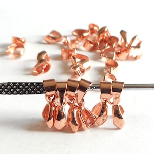 May include: A group of copper-colored metal pendant clasps, some are hanging from a silver tool, others are scattered on a white surface.
