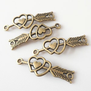 May include: Four bronze-colored metal charms in the shape of Cupid's arrows. Each charm features a heart-shaped arrow head with a smaller heart in the center, a shaft, and a feather fletching. The charms have small loops for attaching to other items.