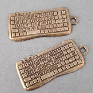 Pode incluir: Dois pingentes de teclado de metal dourado antigo com um layout de teclas e a palavra "SUCCEST" no topo.