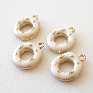 Könnte beinhalten: Vier weiße Donut-Anhänger mit goldenen Akzenten. Die Anhänger sind glasiert und haben Streusel. Sie sind perfekt, um Schmuck oder Bastelarbeiten eine Prise Verspieltheit zu verleihen.