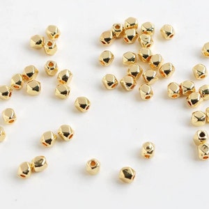 May include: A collection of small, gold-colored, faceted beads scattered on a white surface. Each bead has multiple sides and a hole for stringing. The beads have a shiny, metallic finish, suitable for jewelry making or craft projects.