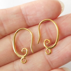May include: Two gold-colored earring hooks. Each hook has a curved, oval shape with a looped bottom and a decorative swirl at the base of the hook. The hooks are designed for jewelry making and are shown against a light background.