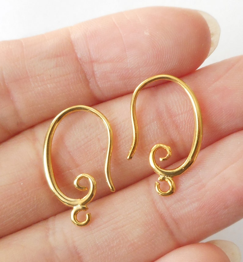 May include: Two gold-colored earring hooks. Each hook has a curved, oval shape with a looped bottom and a decorative swirl at the base of the hook. The hooks are designed for jewelry making and are shown against a light background.