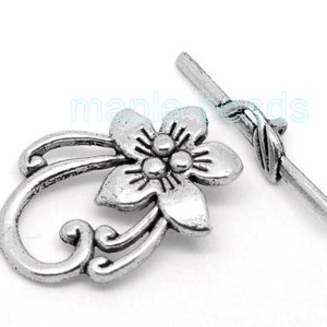 Peut inclure: Un ensemble de fermoir à bascule argenté. Le fermoir présente un motif floral détaillé avec un groupe central de trois petites sphères. La barre de bascule est droite avec un accent en forme de feuille. Le fermoir est probablement destiné à la fabrication de bijoux.