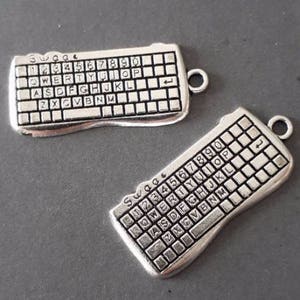 Pode incluir: Dois pingentes de teclado prateados com um design em relevo. Cada pingente tem um layout de teclado padrão com as letras e números impressos nas teclas. Os pingentes têm um pequeno laço na parte superior para prender a um colar ou pulseira.