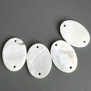 May include: Four oval-shaped, white mother-of-pearl pendants with two holes each. The pendants have a pearlescent sheen and are arranged on a gray surface. These pendants are suitable for jewelry making.