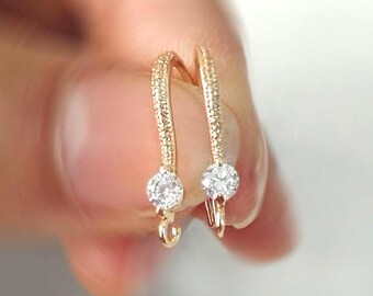 May include: A pair of gold-toned earring hooks featuring a curved design with a textured surface and a single round clear stone set at the base of each hook. Each earring component includes a small loop at the bottom for attaching additional jewelry.