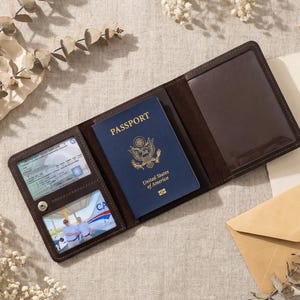 Può includere: Porta passaporto in pelle marrone aperto con un passaporto blu degli Stati Uniti all'interno. Il porta passaporto contiene anche fessure per carte e una tasca trasparente. Una piccola busta marrone e fiori secchi sullo sfondo.