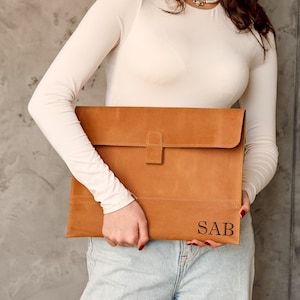 Può includere: Una custodia per laptop in pelle marrone chiaro con chiusura a patta e le iniziali "SAB" incise nell'angolo in basso a destra. La custodia è tenuta da una persona che indossa una camicia bianca a maniche lunghe e jeans chiari.