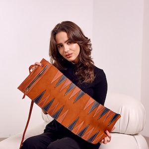 May include: A brown leather backgammon board with a zipper closure and a handle. The board features a geometric design of alternating brown, black, and tan triangles. The board is rectangular and has a leather strap.