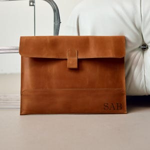 May include: A brown leather laptop sleeve with a flap closure and a tab. The sleeve has a rectangular shape with a stitched design and the initials "SAB" embossed on the lower right corner.