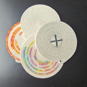 May include: A collection of four circular coasters. The coasters are made of coiled rope, with a natural off-white color. Two coasters have colorful patterned fabric inserts, and one has a dark green cross design.
