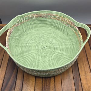 May include: A round, light green woven basket with two handles. The basket has a spiral design on the inside and a decorative rim with pink, white, and tan accents. It sits on a wooden tray.