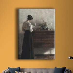Puede incluir: Una pintura enmarcada representa a una mujer con blusa blanca y falda negra, leyendo cerca de un mueble de madera con velas y arreglos florales. La obra está en una pared mostaza sobre un sofá gris.