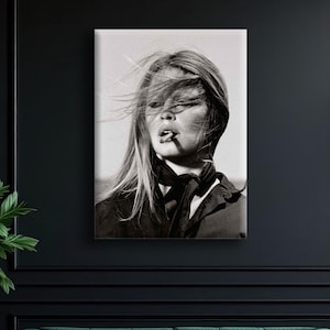 Puede incluir: Retrato en blanco y negro de una mujer con el pelo largo al viento, fumando un cigarrillo. Lleva un top oscuro y una bufanda. La imagen se muestra en un lienzo, montado en una pared oscura sobre un sof&aacute; de terciopelo verde.