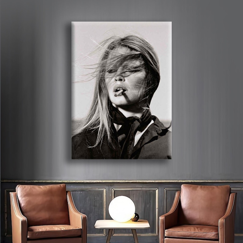 Puede incluir: Retrato en blanco y negro de una mujer con pelo largo y rubio, vestida con un top y una bufanda oscuros, fumando un cigarrillo. La imagen es una impresi&oacute;n sobre lienzo, expuesta en una pared. El viento le despeina el pelo.