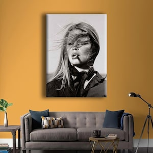 Puede incluir: Retrato en blanco y negro de una mujer fumando un cigarrillo, con el pelo al viento. Lleva una bufanda y una chaqueta oscuras. La imagen es una impresi&oacute;n en lienzo, colgada en una pared mostaza.