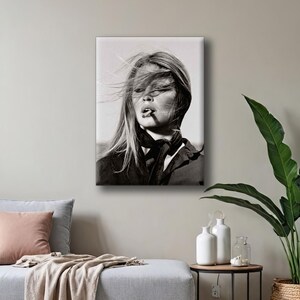 Puede incluir: Impresi&oacute;n en lienzo en blanco y negro de una mujer con el pelo largo al viento, fumando un cigarrillo. Lleva una bufanda y una chaqueta oscuras. La imagen es un retrato que captura un momento de estilo y desaf&iacute;o.