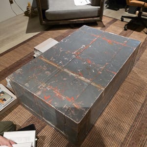 Puede incluir: Una mesa de centro rectangular con un acabado gris desgastado y detalles en rojo. La mesa es de madera y tiene forma de caja. Se encuentra sobre una alfombra tejida en un salón.
