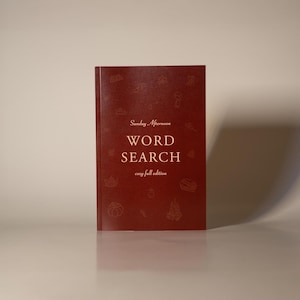 Puede incluir: Un libro "Word Search" de color granate con el texto "Sunday Afternoon" y "cozy fall edition". La portada presenta peque&ntilde;as ilustraciones de color claro. El libro est&aacute; de pie sobre una superficie reflectante.