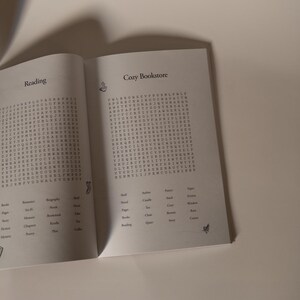 Puede incluir: Un libro abierto con el t&iacute;tulo "Cozy Bookstore" y la palabra "Reading" en la p&aacute;gina izquierda. Las p&aacute;ginas contienen un juego de b&uacute;squeda de palabras. El libro est&aacute; sobre un fondo neutro.