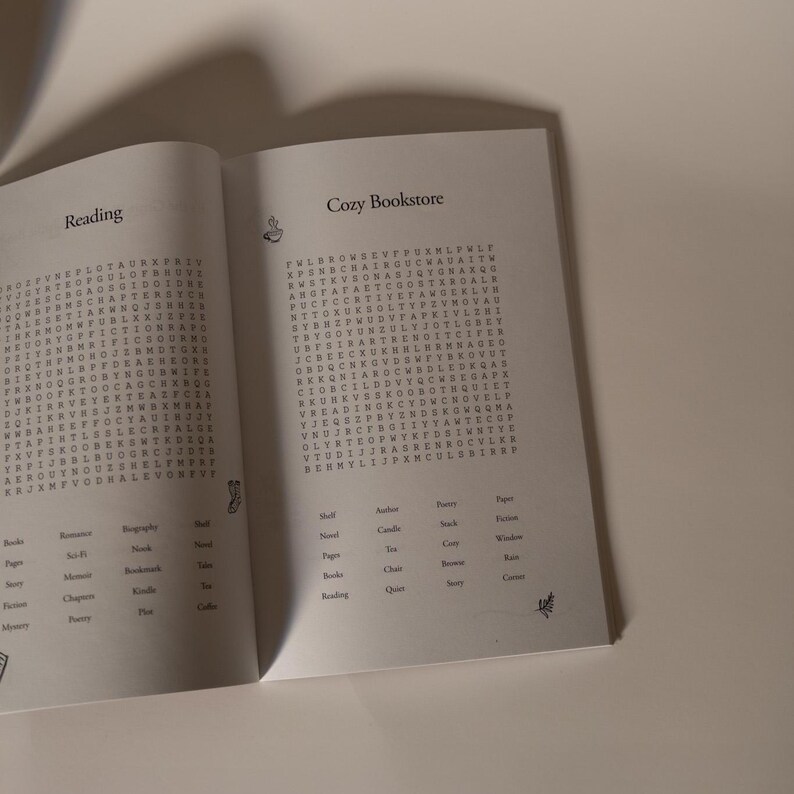 Puede incluir: Un libro abierto con el t&iacute;tulo "Cozy Bookstore" y la palabra "Reading" en la p&aacute;gina izquierda. Las p&aacute;ginas contienen un juego de b&uacute;squeda de palabras. El libro est&aacute; sobre un fondo neutro.