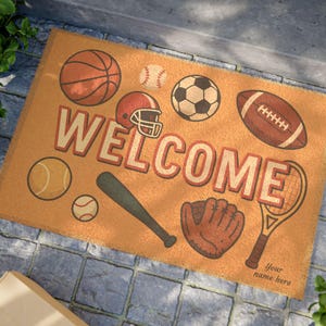 May include: A rectangular doormat with a tan background features illustrations of sports equipment, including a basketball, baseball, and football. The word "WELCOME" is displayed in large, retro lettering. A baseball bat, glove, and tennis racket are also shown.