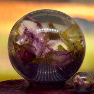 May include: A large clear resin sphere containing dried pink and purple rose petals and green leaves, accompanied by a smaller matching resin sphere. The spheres have a smooth, glossy finish and are positioned to show the floral inclusions clearly.