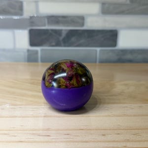 May include: A spherical paperweight featuring a clear resin top half embedded with dried pink and green rosebuds, and a solid opaque purple resin bottom half. The object has a smooth, glossy finish and a rounded shape, displaying floral elements.