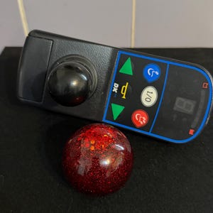 May include: A black wheelchair joystick controller featuring a central black knob, a digital display, and buttons with green, blue, red, and white icons, accompanied by a spherical red resin topper filled with fine red and orange glitter.